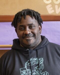 Headshot of Greg, a black man with short hair in dreads wearing a black graphic hoodie, smiling