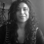 A black-and-white picture of Maria, a Latina woman with long curly hair, in front of a bookshelf with lamp