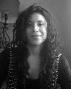 A black-and-white picture of Maria, a Latina woman with long curly hair, in front of a bookshelf with lamp