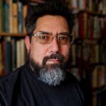 Headshot of Adam Collet, a man with rectangular glasses, dark hair, and a beard, in front of bookshelf