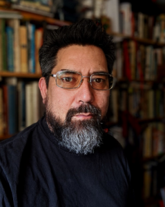 Headshot of Adam Collet, a man with rectangular glasses, dark hair, and a beard, in front of bookshelf