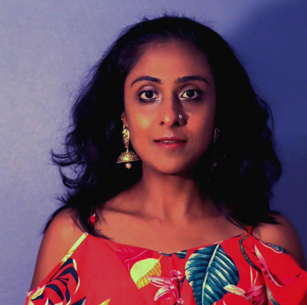 Teaching Artist Divya stands in front of a plain background with side lighting. She wears a red top and gold earrings.