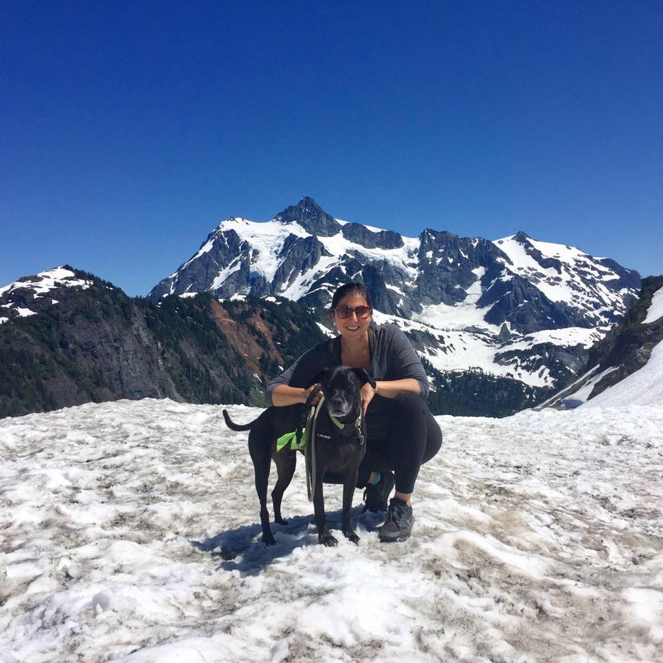 A woman poses with her black dog at on snowy mountain close to the peak.