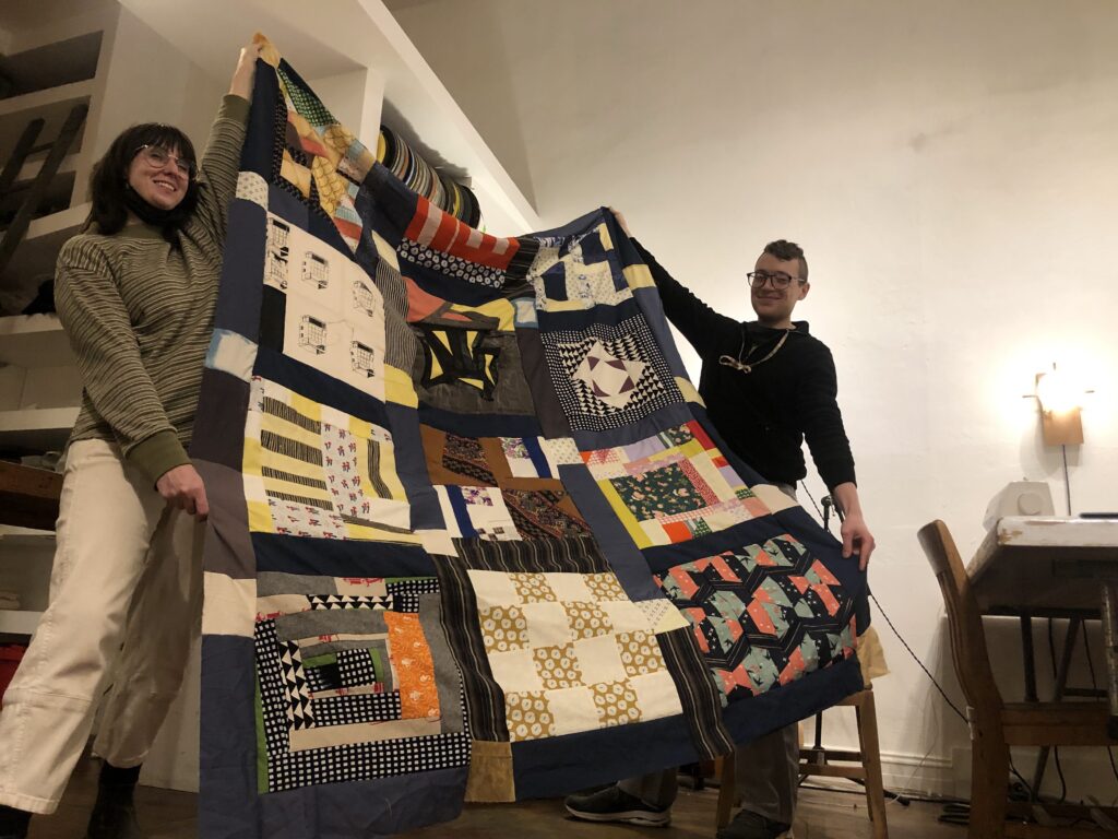 Artists Dani Hopple and Sander Moberg holding up a quilt composed of non-matching squares and a blue border