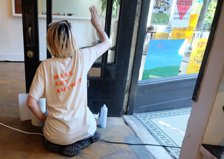 A youth wearing a shirt reading "Make Art Anyway" sitting in front of a doorway, cleaning a glass door