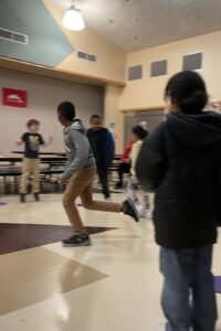 An elementary student in focus while he runs in a school cafeteria, in a circle of peers