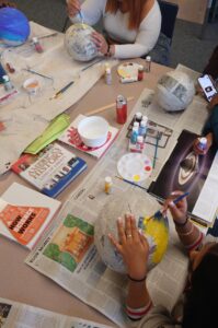 Students paint paper mache spheres, with a magazine open to a photo of Saturn between them