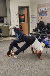 An elementary student mirrors a teacher in a triangular pose, holding head against floor, with one leg raised