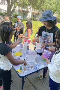 Teaching artist a white table full of supplies to make slime, talking with youth around them, outside