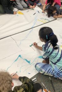 Elementary students draw on a big piece of white paper on the floor, with nature as a theme