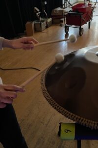 Hands playing a pan drum with mallets in a room with wooden floors