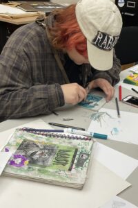 A student carves out a stamp block that has been colored blue, a sketchbook next to them with drawings on cover