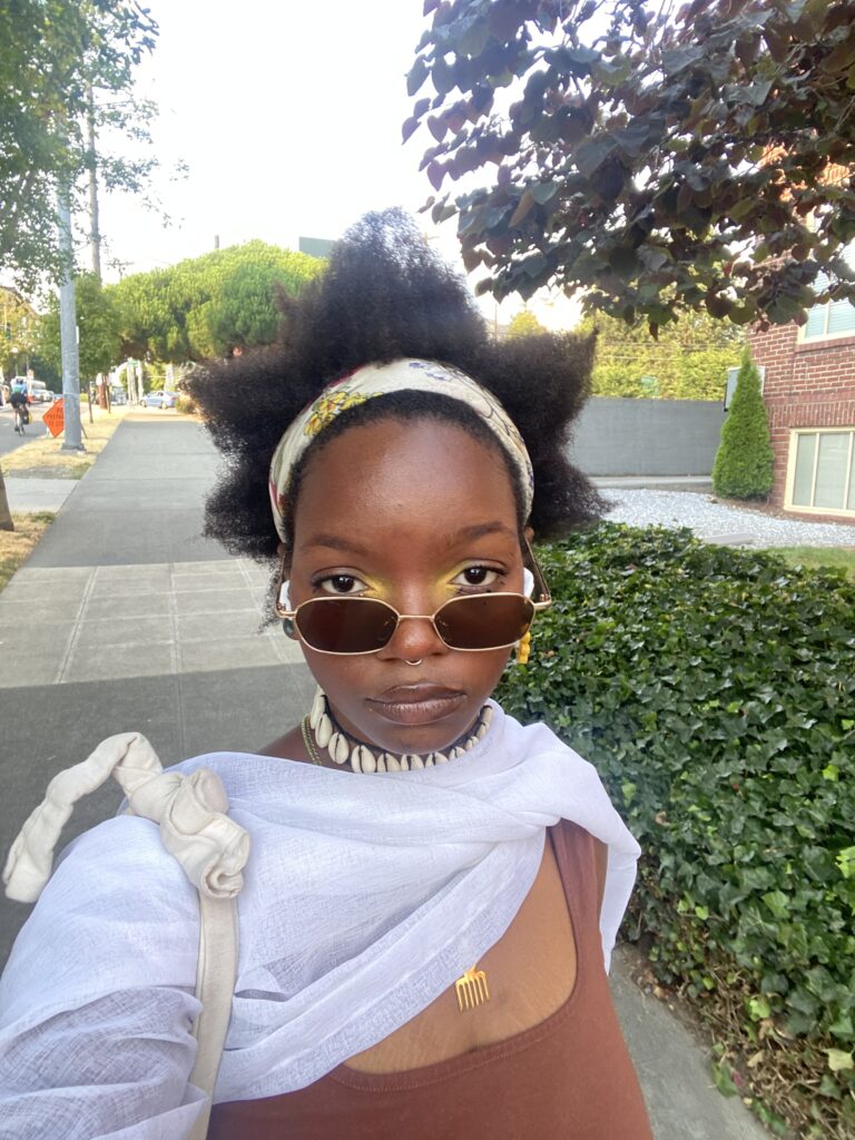 A selfie of Yaz, a black person in their 20s on a sidewalk, wearing white sunglasses and their shaped in 5 points