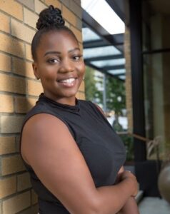 Headshot of Antonesha, a black woman with hair in a top bun, posing against a brick wall, smiling