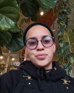 A selfie of Arielle, a Latine human wearing a black beanie, hoodie, and glasses, with plants in the background