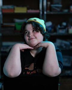 A headshot of Eris, a white woman in her 20s with short hair and a beanie, with her hands framing her face