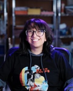 A headshot of Grecia, a Latina woman in her 20s wearing an Arts Corps shirt and glasses, with shelves as a blurred background