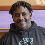 Headshot of Greg, a black man with short hair in dreads wearing a black graphic hoodie, smiling