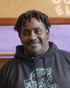 Headshot of Greg, a black man with short hair in dreads wearing a black graphic hoodie, smiling