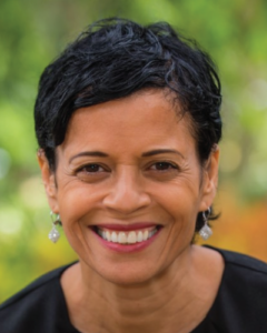 Headshot of Shawn, a black woman with short hair, smiling against blurred greenery, wearing a black top and earrings