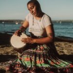 Photo of Taylor, a black person, drumming on a rock next to the ocean, wearing a skirt and long beaded earrings