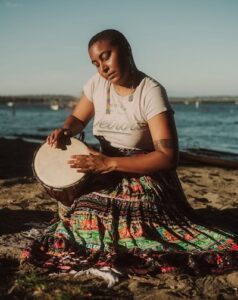 Photo of Taylor, a black person, drumming on a rock next to the ocean, wearing a skirt and long beaded earrings