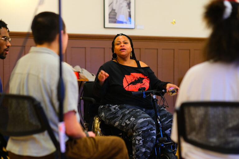 Teaching Artist Erica in focus seated in a circle of young adults, singing and gesturing with her hand