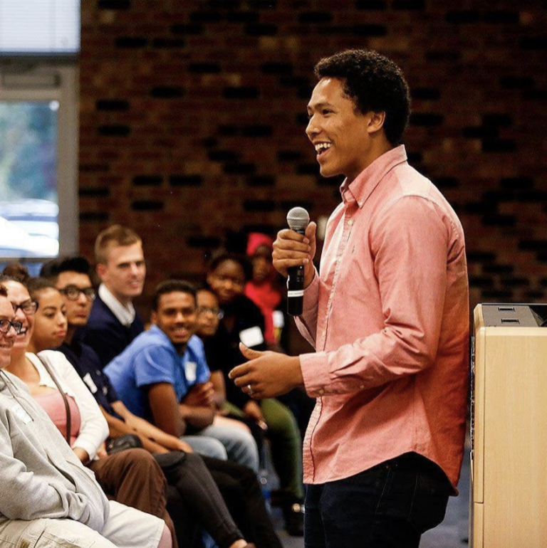 Christian Paige, a young black man wearing a salmon button up, holding up a mic and smiling in front of a seated crowd