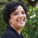A headshot of Sue, a Latine person with short curly hair smiling at the camera, a tree in the background.