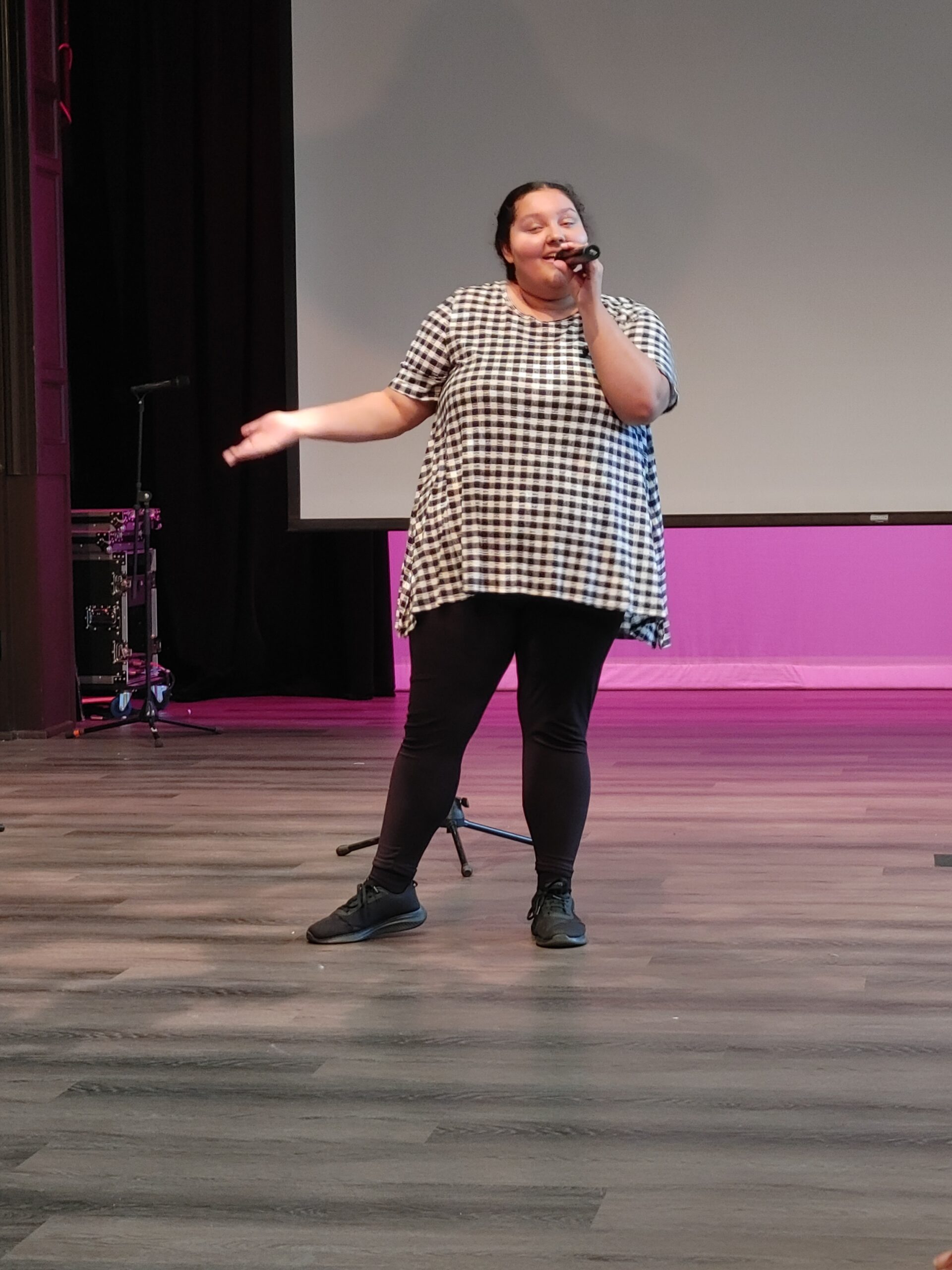 Photo of Jordan Johnson, a woman in her twenties with curly hair pulled back, wearing a checkered shirt, singing into a mic.