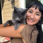Photo of Tianna Eddy, a Mexican-Japanese woman in her early twenties, smiling over her shoulder while carrying a grey cat