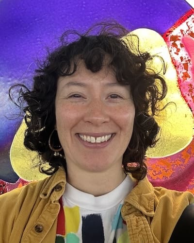 Alyza DelPan-Monley Headshot of a middle aged person with a wide smile and curly hair, against a colorful background.