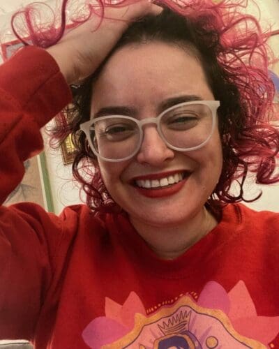 Headshot of Ana María, a Mexican woman with red curly hair and glasses, wearing a red hoodie, smiling