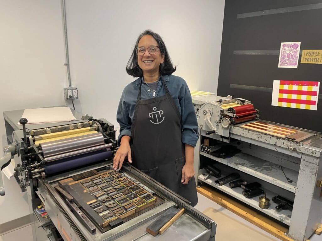 Picture of Chaya Keefe, an Indian woman with shoulder lenght hair, smiling behind a letter press with wooden blocks.