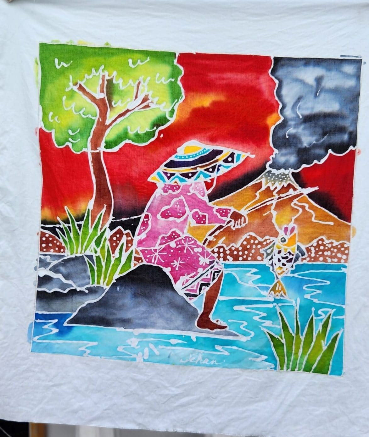 A Batik tapestry of a person sitting on a rock, fishing, with a tree, a volcano, and a red sky behind them