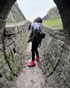 Jéhan, a black person wearing pink boots and a white scarf, in the middle of a brick-lined road beneath an arched bridge.