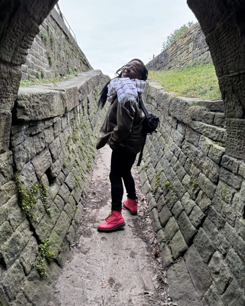 Jéhan, a black person wearing pink boots and a white scarf, in the middle of a brick-lined road beneath an arched bridge.