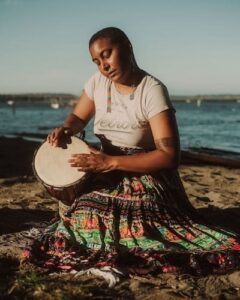 Photo of Taylor, a black person, drumming on a rock next to the ocean, wearing a skirt and long beaded earrings
